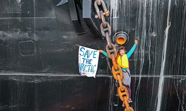 A young woman hanging off of a thick anchor chain raises her hands in a gesture of triumph. She's holding a sign that says "Save the Arctic."