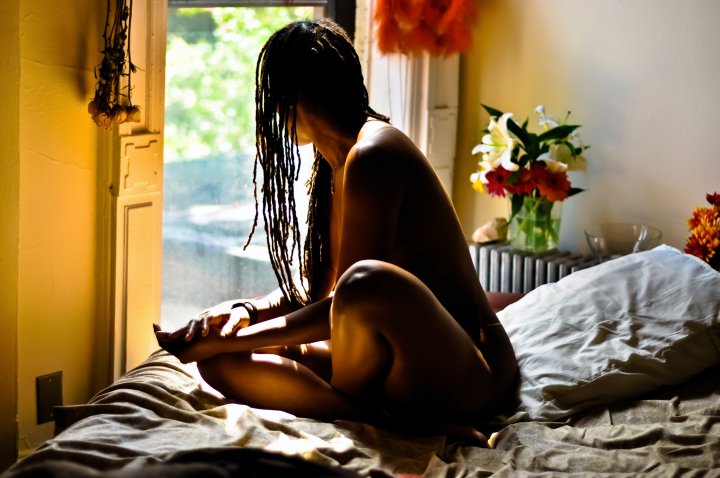 naked african-american woman sitting on a bed, looking out a window
