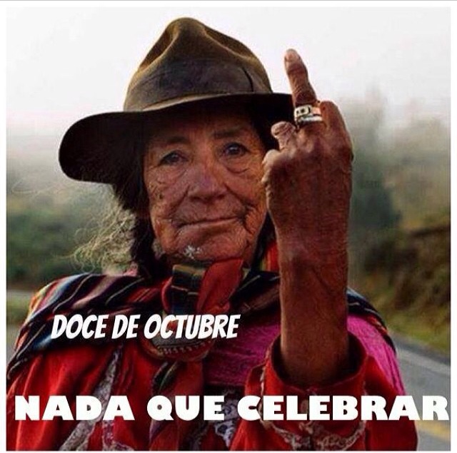An indigenous woman wearing red clothing and a felt hat flips off the camera. Image reads: "The 12th of October is nothing to celebrate!"