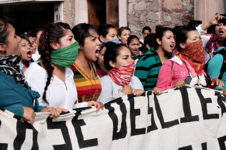 A line of young women holding a banner, their mouths open in protest. They are wearing bandanas on their faces, and many of them have their hair braided.