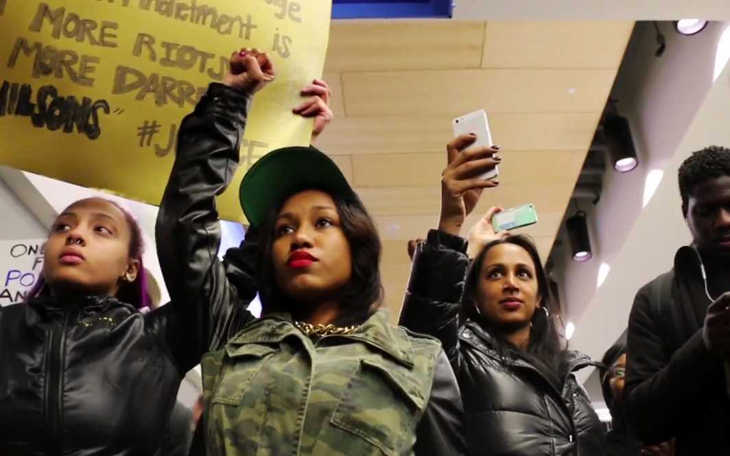 Black women with hands raised in fists, cell phones up documenting protests, holding signs. The woman in the center is a femme in bold red lips.
