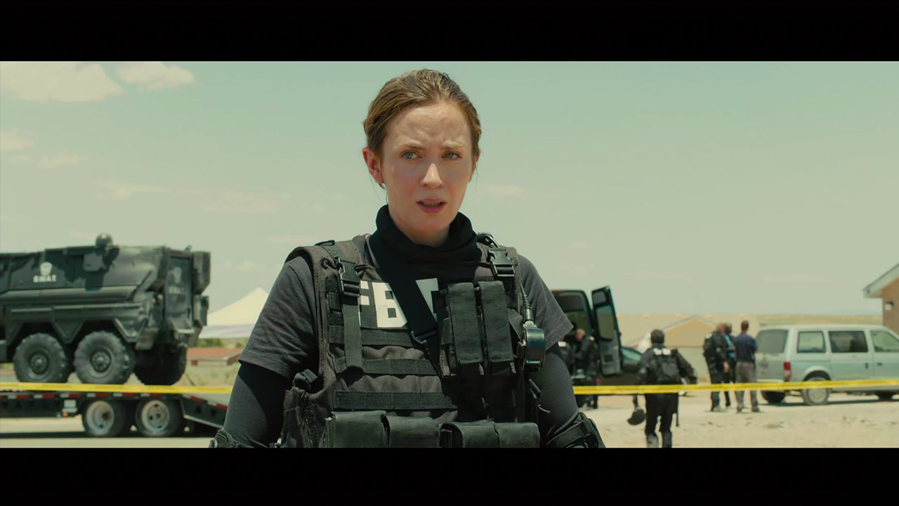 Actress Emily Blunt, woman with dark hair and fair skin standing in the open air of a crime scene in the desert, and wearing FBI gear