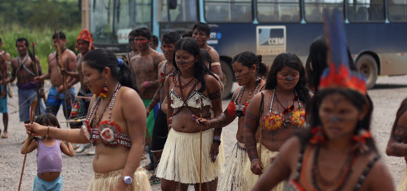 Indigenous women from the Xingu River walk wearing traditional grass skirts.
