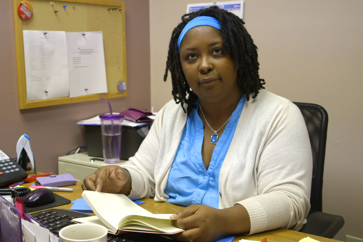 A black woman wearing scrubs, with locs in a headband, looks at the camera while at her desk. She is an abortion provider.