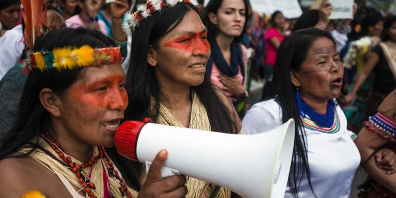 Three indigenous women walk, arms linked, faces painted. One of them is holding a megaphone.