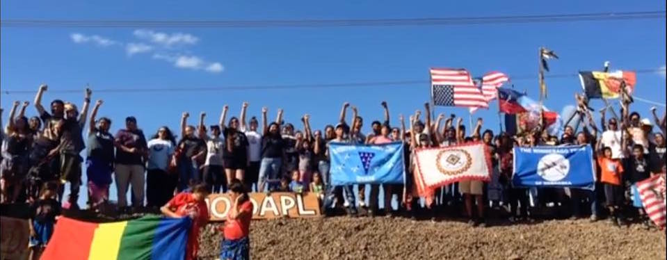 A group of 50+ protestors stand on a mound of dirt holding flags, their fists in the air.