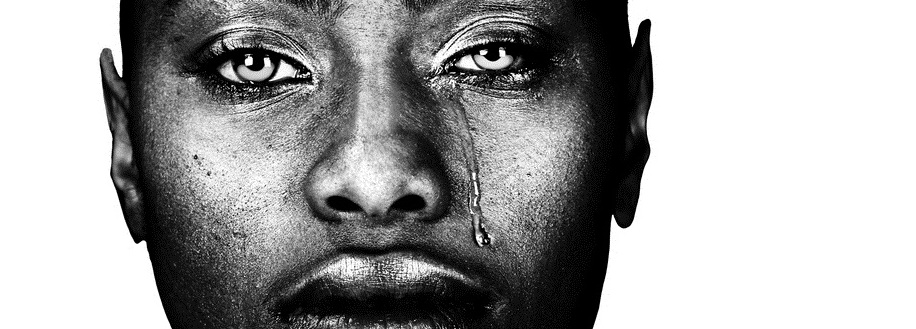 A black woman with dark skin looks at the camera. A tear drips down her cheek.