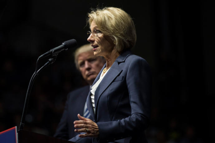 Betsy Devos stands at a mic with Donald Trump behind her.