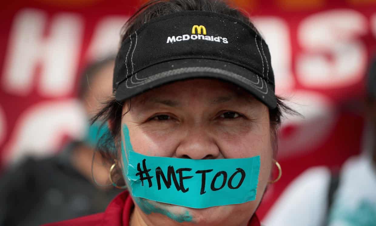 A woman participating in yesterday's McDonald's strike wears a baseball hat looks directly at the camera. There is a piece of blue tape over her mouth that says #MeToo. She has a blue handprint painted on the left side of her face.