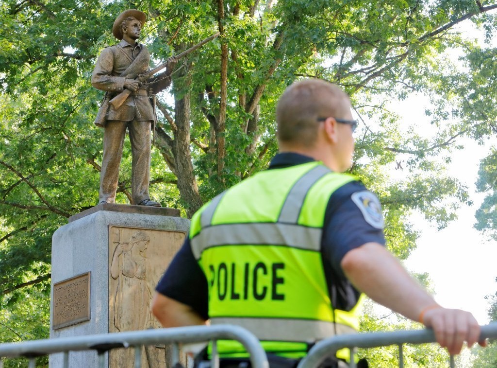 picture of a white police officer in a bright green vest that says "police" on the back standing in front of a metal barricade, facing away from the camera. Behind him is the Silent Sam statue at the University of North Carolina Chapel Hill campus